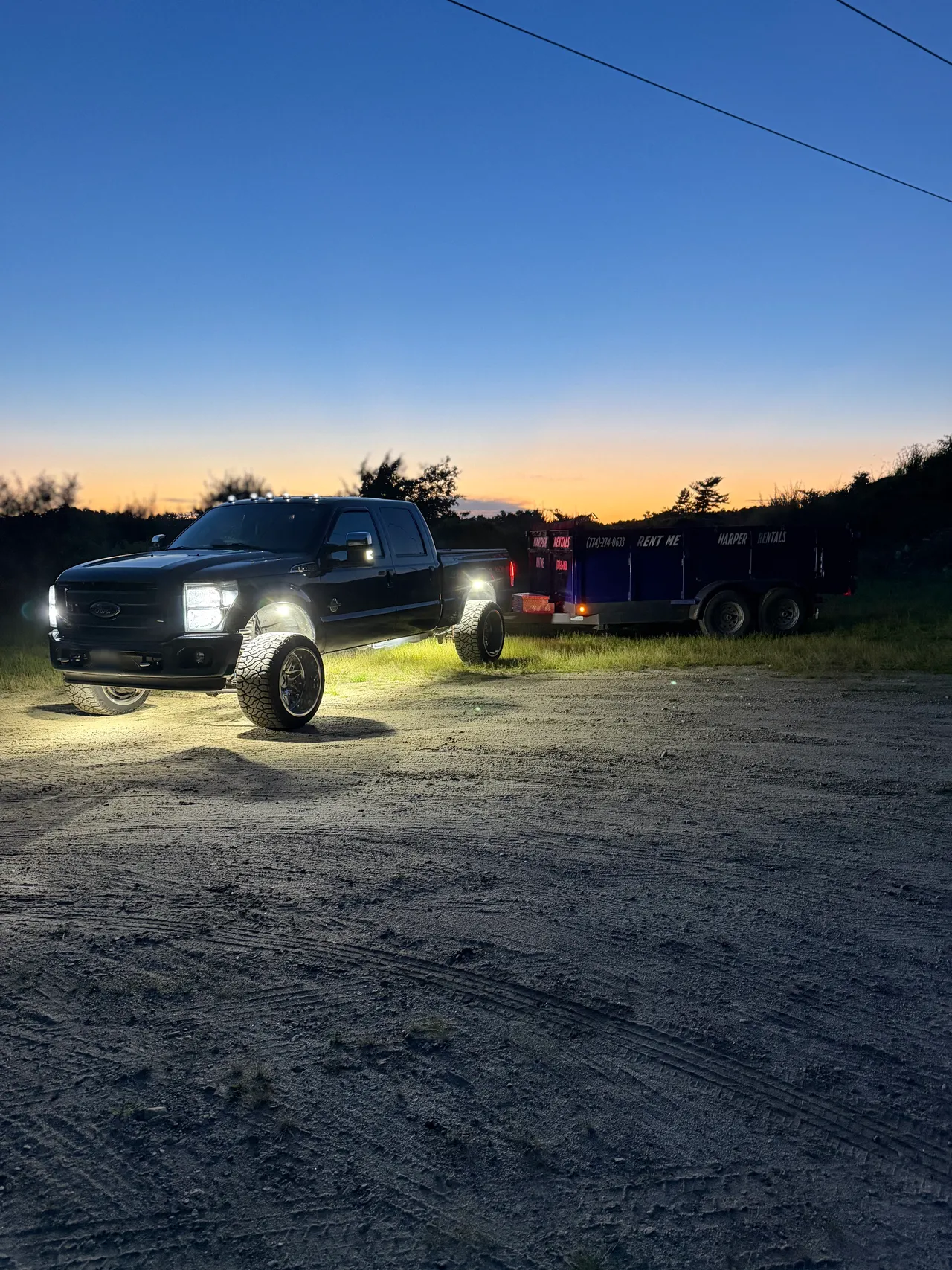 The Harper Rentals pickup at dusk with its headlights and cab lights on, hitched to the company dump trailer on a dirt lot, the trailer lettered RENT ME with the phone number along its side, against a sunset treeline.