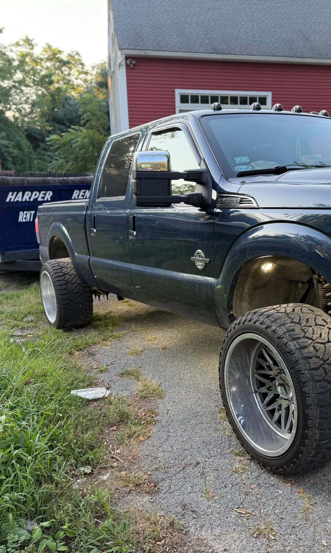 A navy Ford pickup on a gravel drive with the Harper Rentals dump trailer hitched behind it, tarp rolled back along the top rail, parked in front of a red barn.